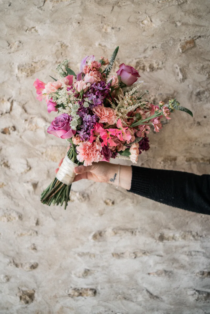 Bouquet de mariée tenu à bout de bras devant un mur de pierre