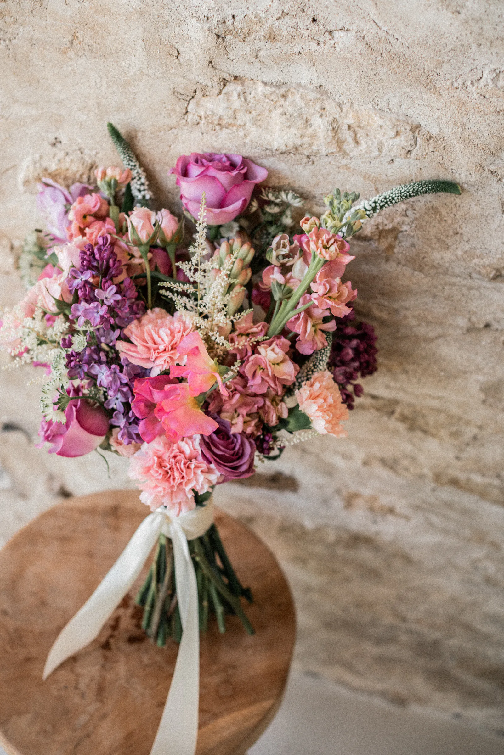 Bouquet de mariée aux teintes rose et violine posé sur un tabouret en bois devant un mur de pierre