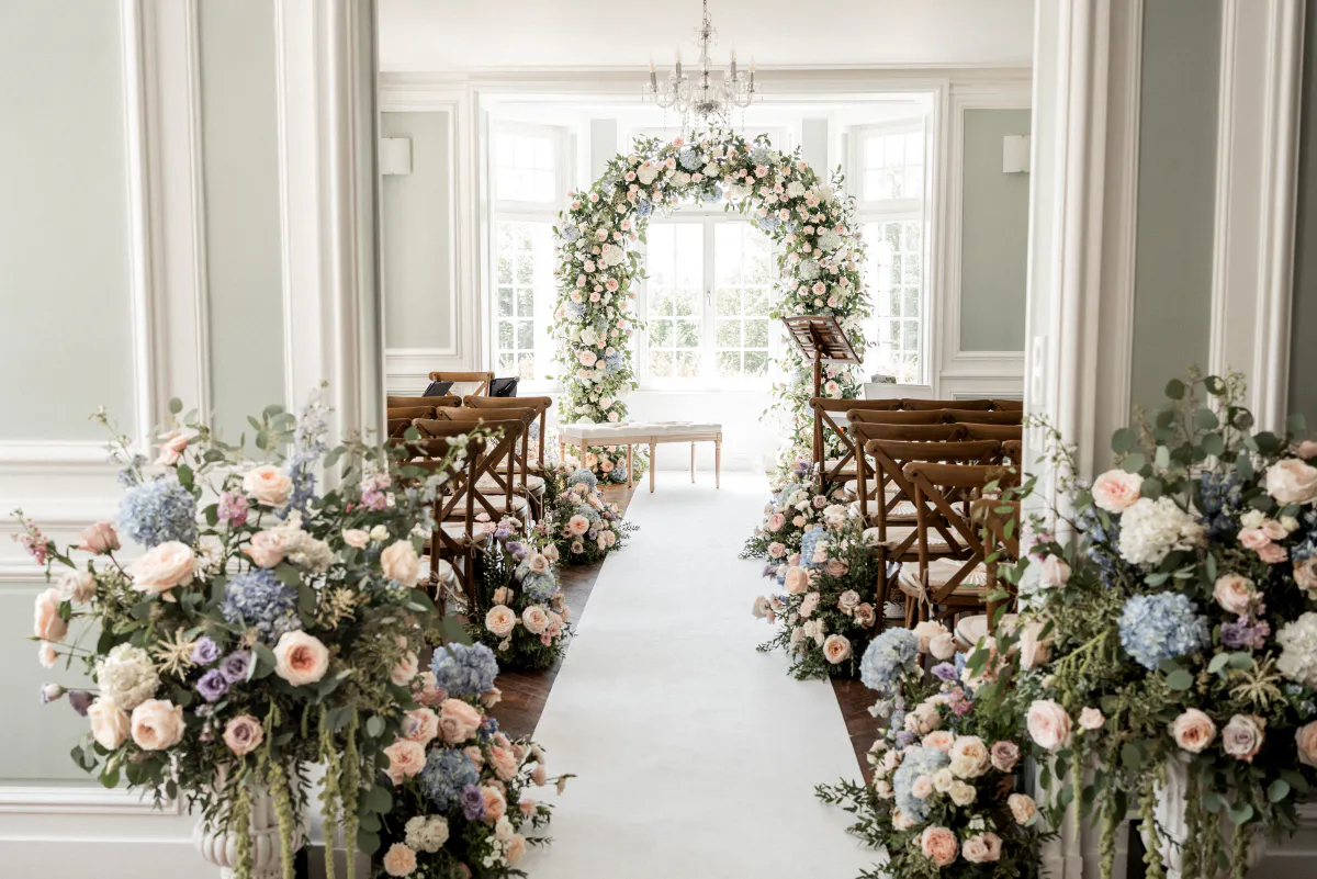 Allée de cérémonie bordée de fleurs menant à une arche florale dans un salon lumineux