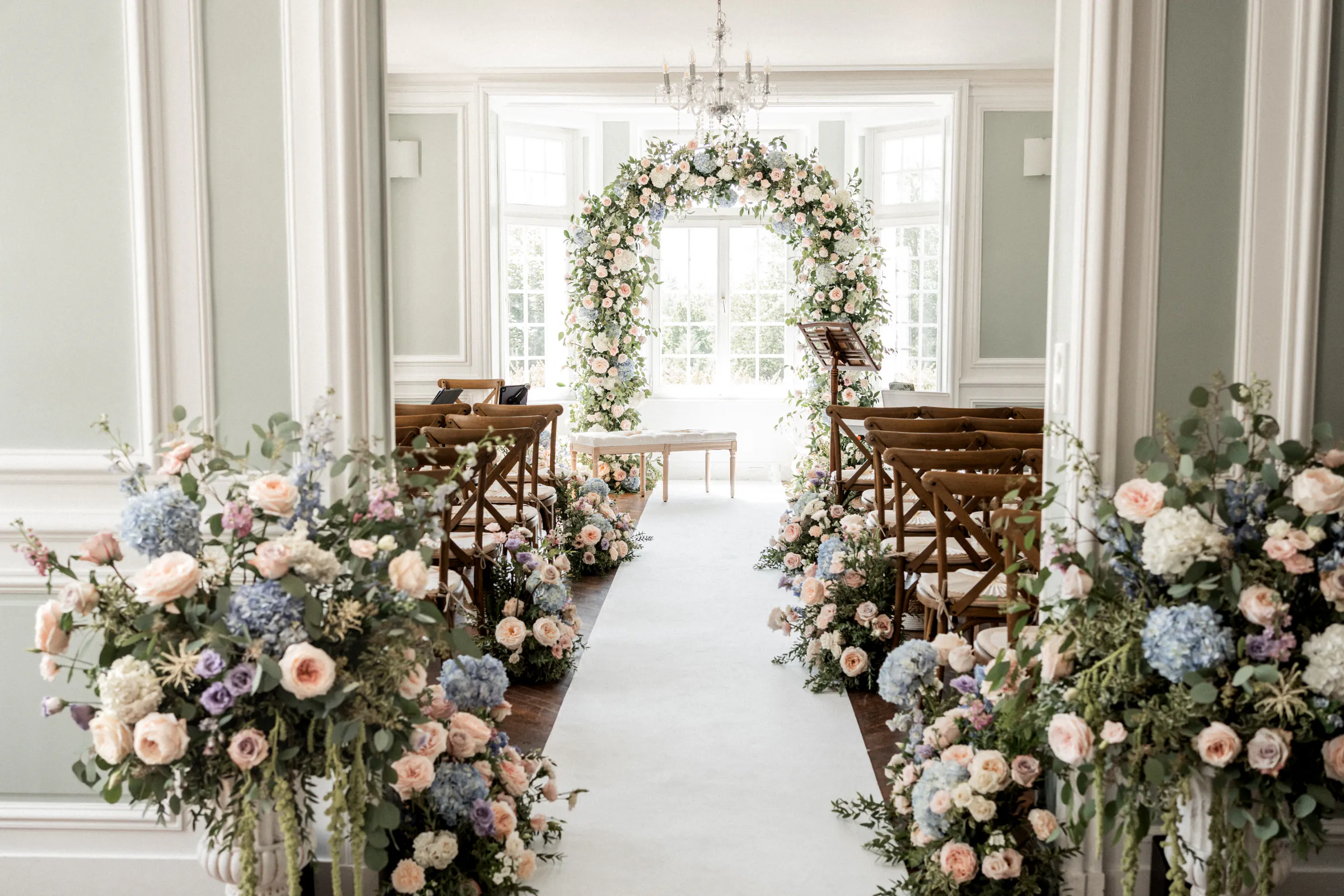 Allée de cérémonie bordée de fleurs menant à une arche florale dans un salon lumineux