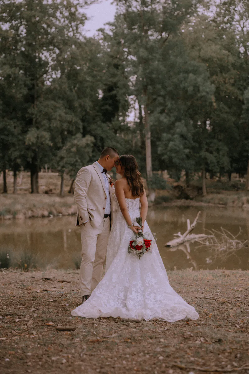 Mariés enlacés au bord d'un étang, la mariée tenant un bouquet de roses rouges