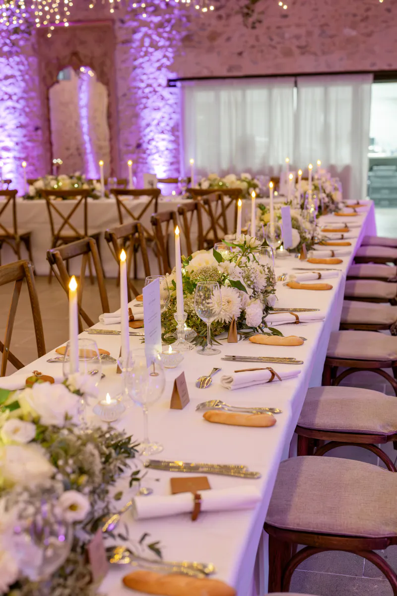 Longue table dressée de bouquets blancs et de bougies sous un éclairage mauve
